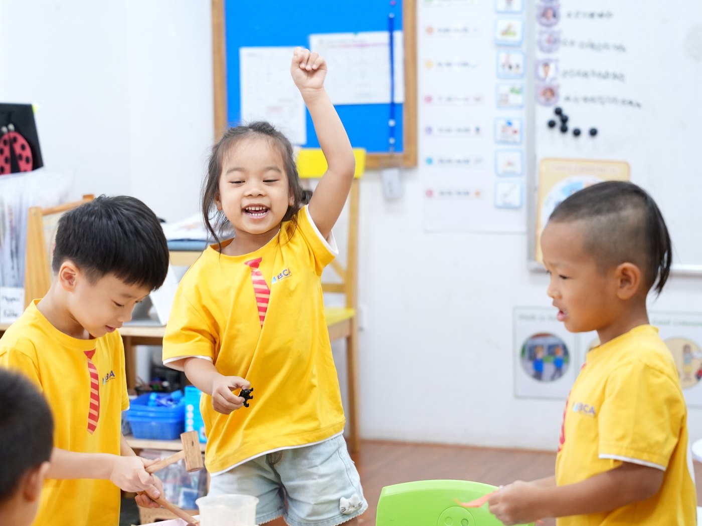 Children using small wooden hammers at a tray while one girl raises her arm and laughs